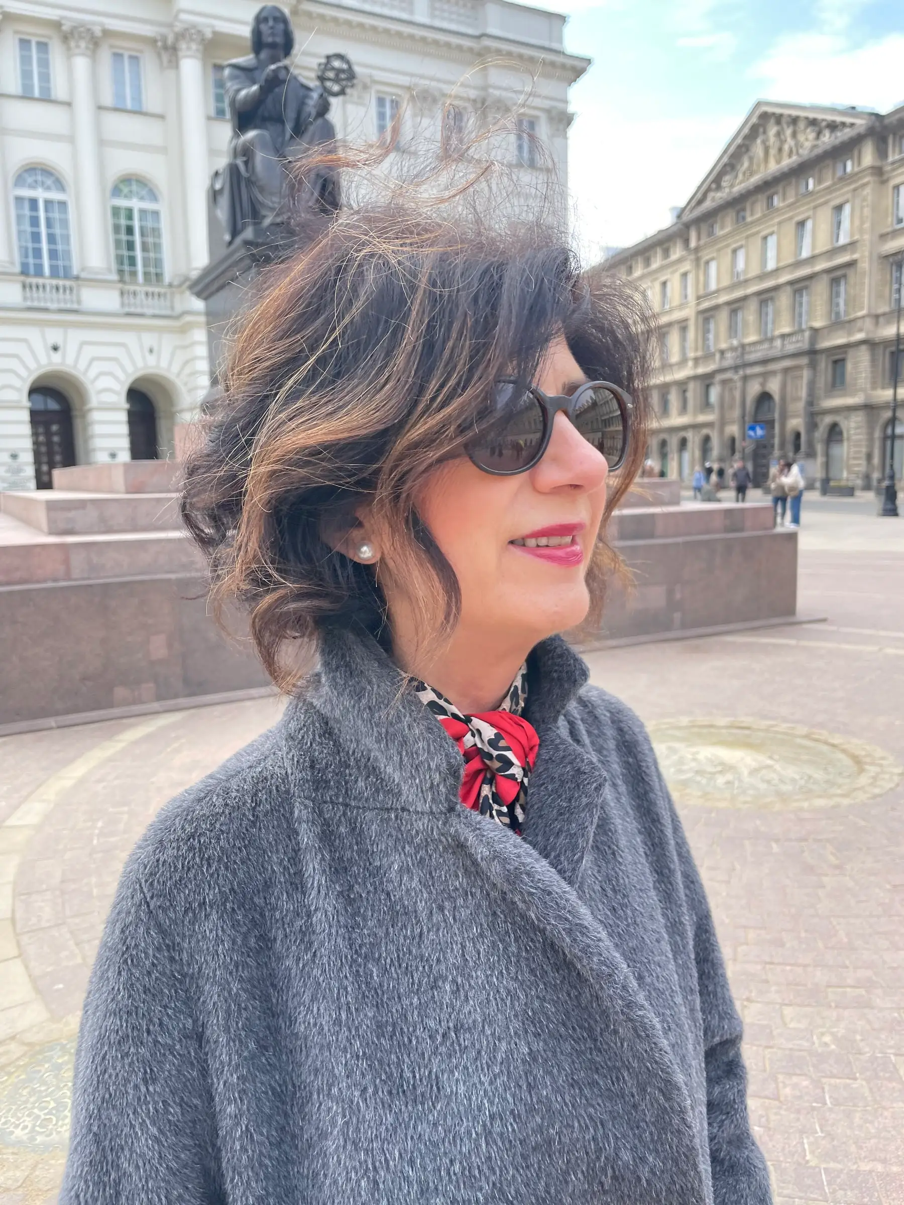 Carmen Díez Grenabuena con abrigo gris, pañuelo rojo y gafas de sol en una plaza, con una estatua y edificios clásicos al fondo